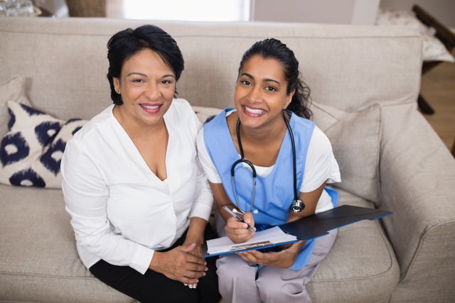 Portrait of smiling patient sitting with doctor checking medical report on sofa at home