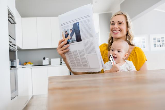 Mother reading newspaper with baby boy in kitchen at home