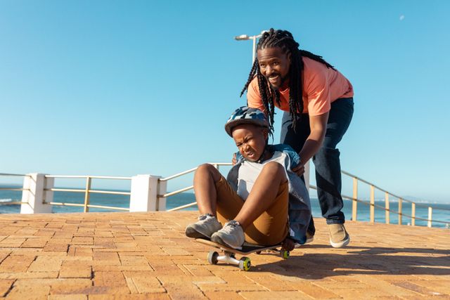 Happy african american father supporting son skateboarding on promenade at sunny day. unaltered, parenting, family, lifestyle and togetherness concept.