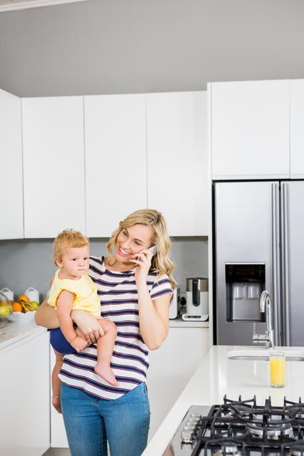 Mother talking on mobile phone while holding her baby girl in kitchen - Download Free Stock Photos Pikwizard.com