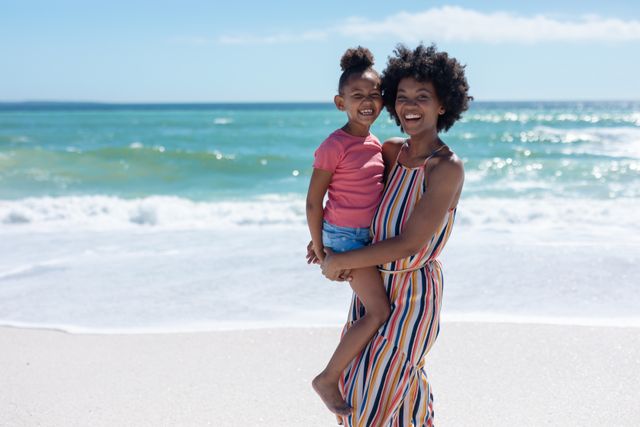 Portrait of smiling african american mother carrying daughter standing at beach on sunny day - Download Free Stock Photos Pikwizard.com