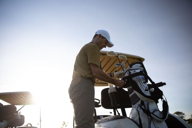Caucasian male golfer practicing on a golf course on a sunny day wearing a cap and golf clothes, taking his golf bag out of a golf cart. Hobby healthy lifestyle leisure.