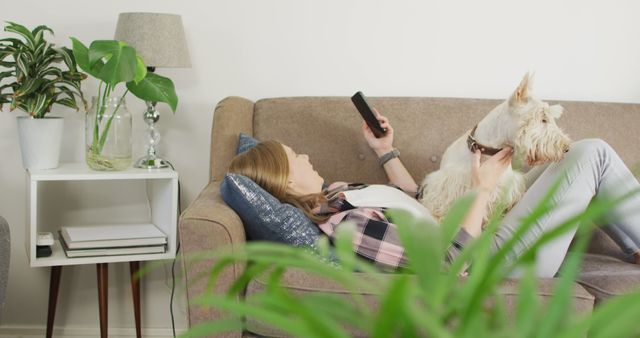 Caucasian woman using smartphone while lying on the couch with her dog at home. pet love and care concept