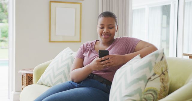 Happy mixed race woman sitting on sofa, using smartphone in living room. domestic lifestyle, enjoying leisure time at home with technology.