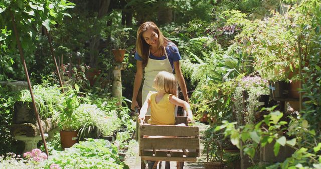 Happy caucasian mother and daughter walking with wheelbarrow in sunny garden. Family, nature, gardening and hobbies.