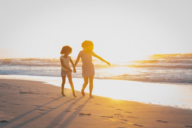 African american mother and daughter holding hands walking on beach at sunset. summer beach vacation by the sea.