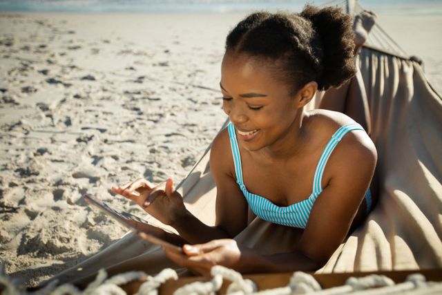 Smiling african american woman sitting in hammock using tablet on sunny the beach by the sea - Download Free Stock Photos Pikwizard.com