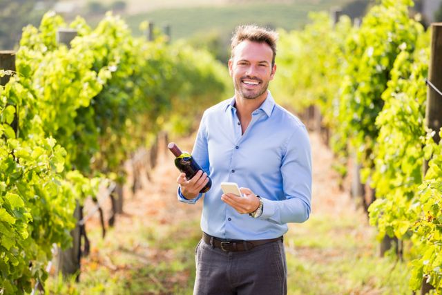 Portrait of smiling man with wine bottle using phone at vineyard on sunny day