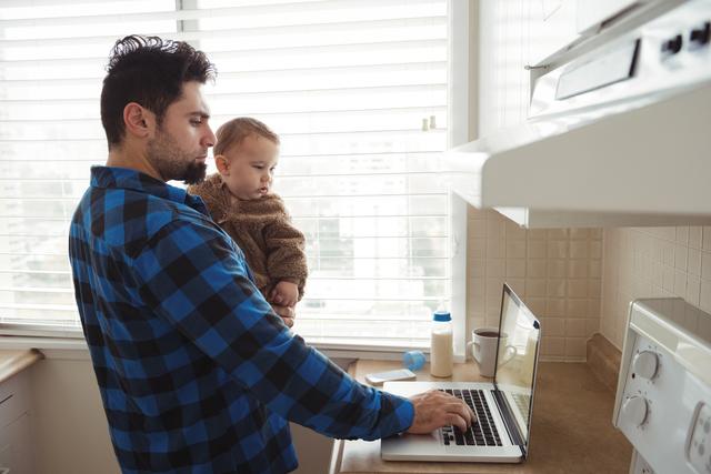 Father using laptop while holding his baby in kitchen - Download Free Stock Photos Pikwizard.com
