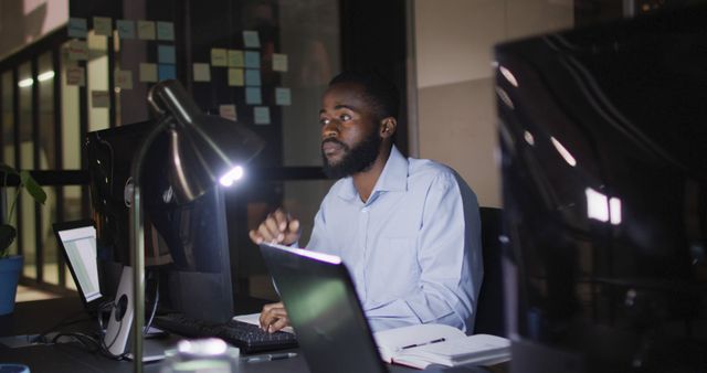 Image of african american businessman sitting at desk using computer at night in office. Business, communication, inclusivity and flexible working concept digitally generated image.