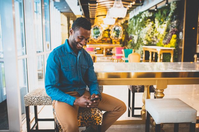 African american man sitting in a cafe using a smartphone and smiling - Download Free Stock Photos Pikwizard.com
