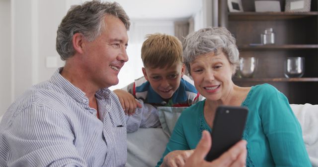 Happy caucasian grandparents and grandson sitting on sofa, using smartphone. Lifestyle, domestic life, communication, family, and togetherness.