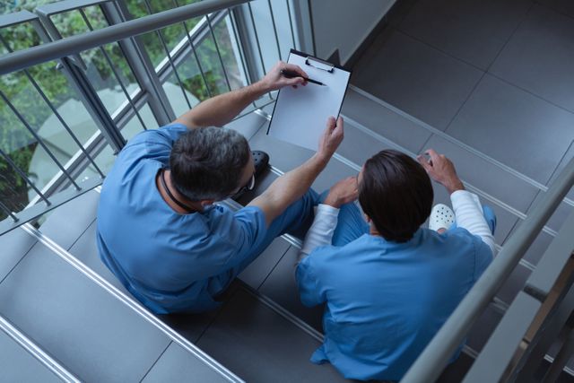 Male surgeons discussing over clipboard while sitting on stairs at hospital - Download Free Stock Photos Pikwizard.com