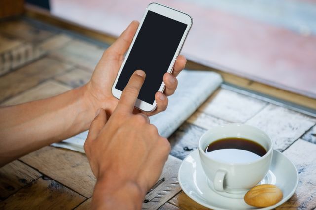 Cropped image of woman holding mobile phone by coffee cup at table