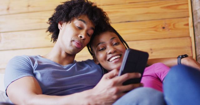 Happy african american couple using smartphone, sitting on sofa in log cabin, slow motion. Domestic life, countryside and nature concept.
