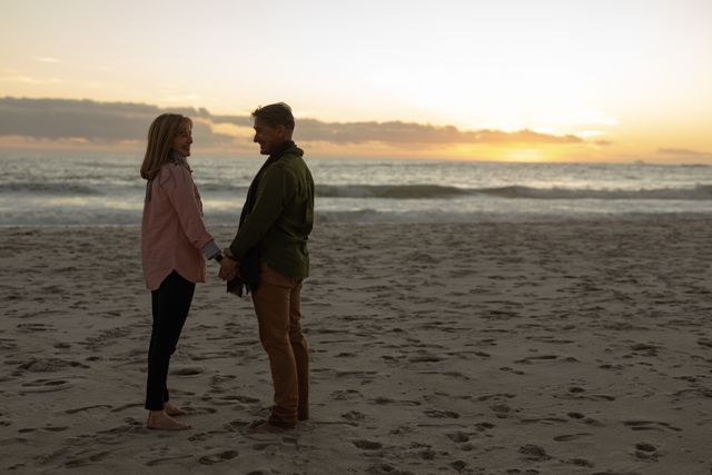 Side view of a happy senior Caucasian couple in love enjoying time in nature together, standing on a beach barefoot, holding hands looking at each other with sea and sunset in the background.