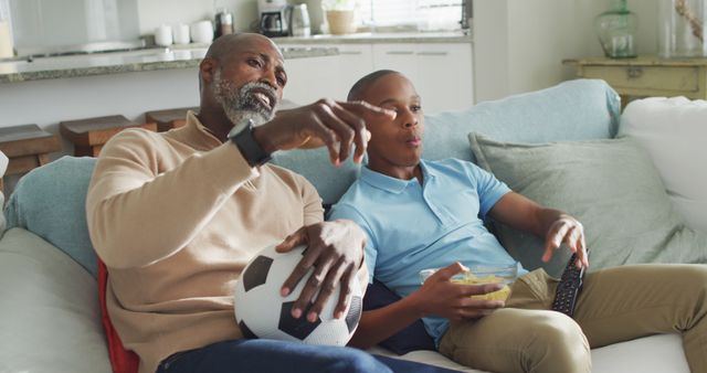 Image of happy african american father and son eating chips and watching tv together. Family, spending quality time together at home.