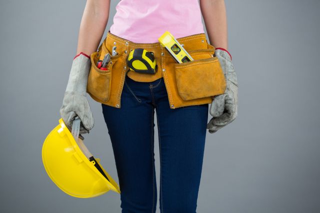 Woman with tool belt and holding hard hat against grey background - Download Free Stock Photos Pikwizard.com
