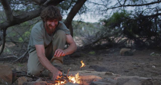 Happy bearded caucasian male survivalist adding kindling to fire at camp in wilderness. exploration, travel and adventure, survivalist in nature.
