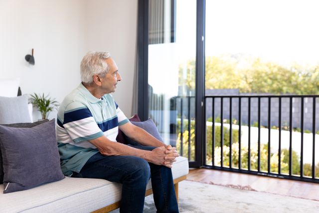 Caucasian senior man contemplating and looking through window while relaxing on sofa in living room. Thoughtful, copy space, unaltered, lifestyle, retirement and home concept.