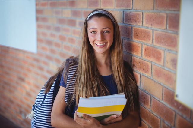 Happy schoolgirl holding books and standing near brick wall - Download Free Stock Photos Pikwizard.com