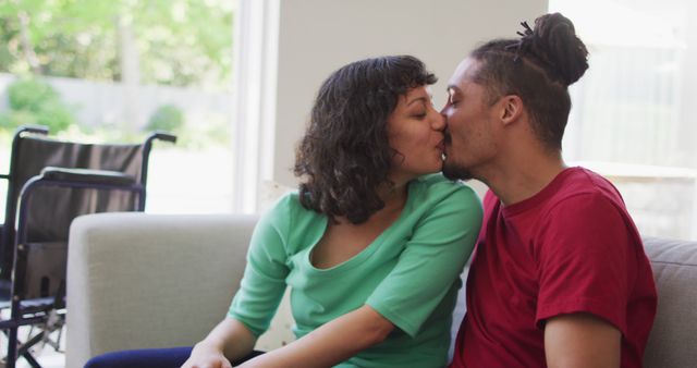 Happy biracial couple sitting on couch kissing in living room, with wheelchair in background. wellbeing and domestic lifestyle with physical disability.