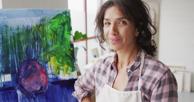 Image of happy biracial female artist posing with painting in studio. Art, crafts, creativity and creation process concept.