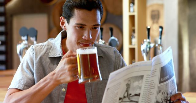 A young Asian man enjoys a pint of beer while reading a newspaper at a bar, with copy space - Download Free Stock Photos Pikwizard.com