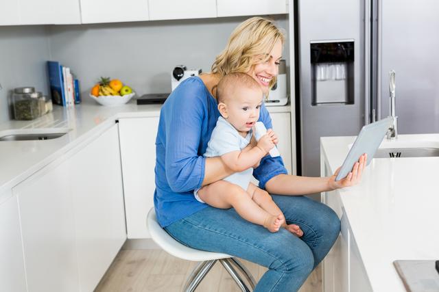 Mother holding baby boy while using digital tablet in kitchen - Download Free Stock Photos Pikwizard.com