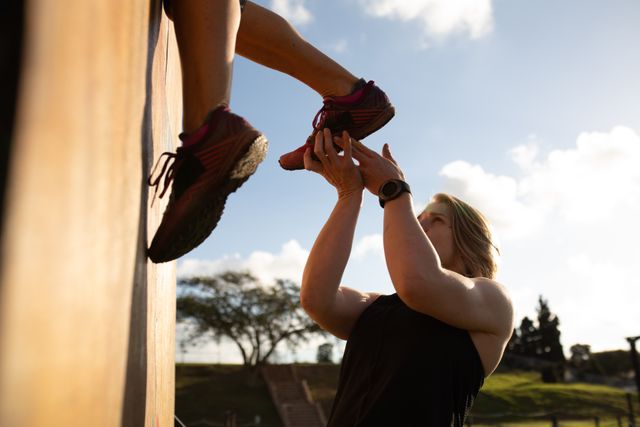 Side view of a Caucasian woman helping another woman climb over a wall, holding her foot to give her a leg up at an outdoor gym during a bootcamp training session
