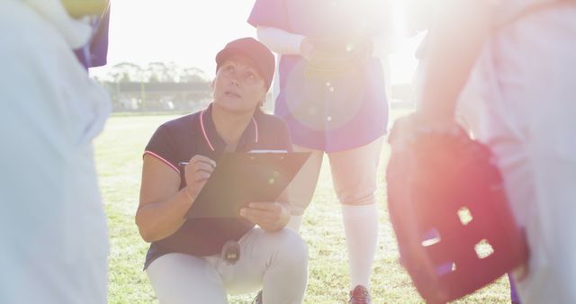 Caucasian female baseball coach squatting writing on clipboard and talking to team on pitch. female baseball team, sports training and game tactics. u