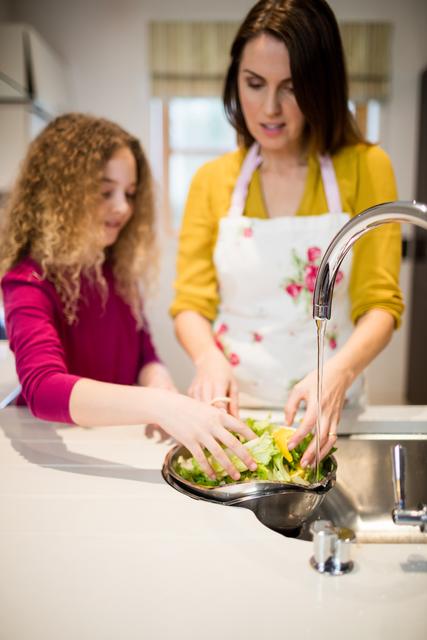 Mother assisting daughter in washing lettuce in kitchen at home