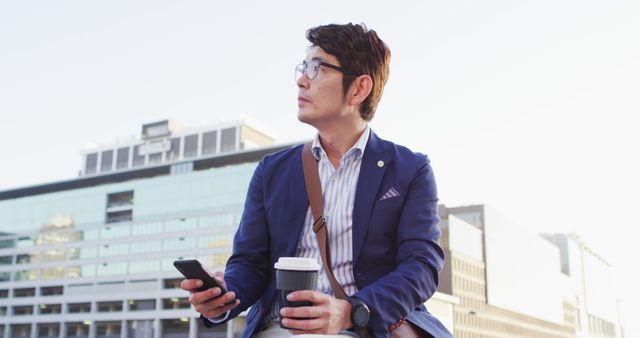 Asian man holding coffee cup and smartphone while sitting on the roof of corporate park. business and lifestyle concept
