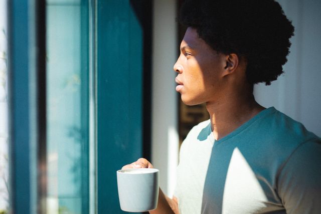 Thoughtful african american young man holding coffee cup while standing by window at home - Download Free Stock Photos Pikwizard.com