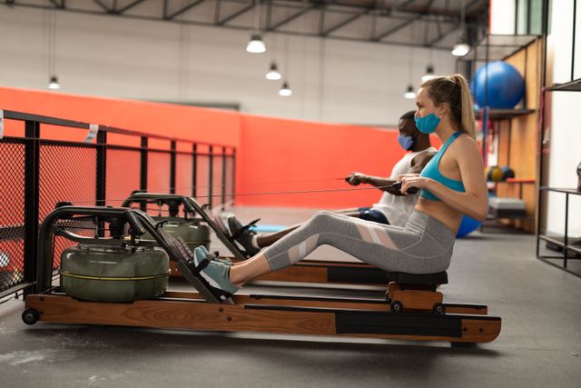 Caucasian woman and an african american man working out on the rolling seat exercise machine. both of them are wearing facemasks.