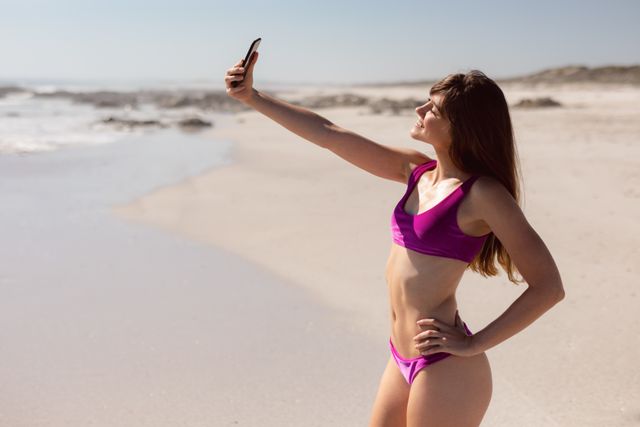 Beautiful young woman in bikini taking selfie with mobile phone at beach in the sunshine