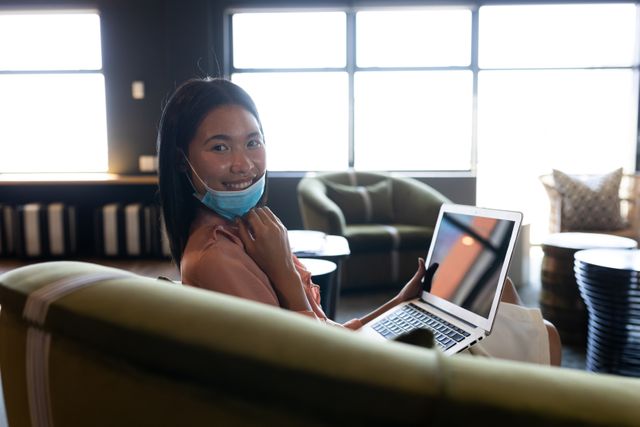 Asian woman wearing a face mask sitting on couch using laptop looking at camera smiling - Download Free Stock Photos Pikwizard.com