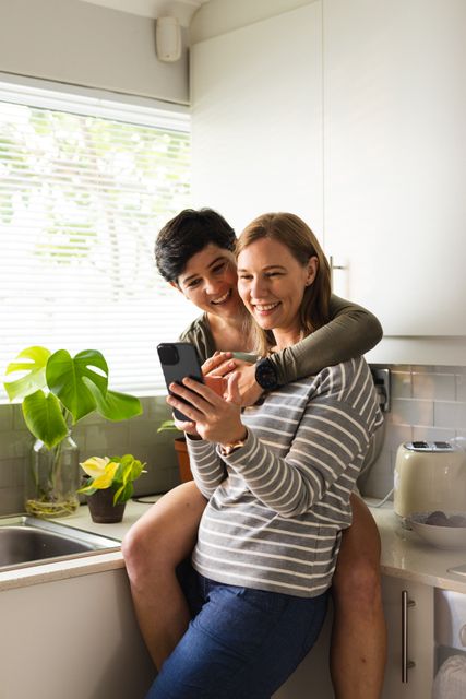 Happy caucasian lesbian woman cuddling girlfriend using smartphone while having coffee in kitchen. Copy space, technology, drink, unaltered, love, togetherness, homosexual, lifestyle and home concept.