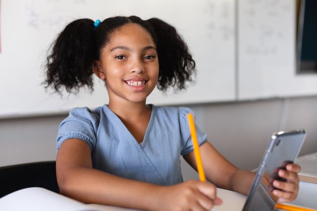 Portrait of smiling biracial elementary schoolgirl with digital tablet sitting at desk in classroom - Download Free Stock Photos Pikwizard.com