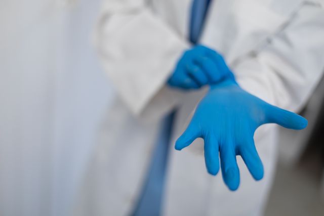 Midsection of doctor in lab coat putting on blue surgical gloves. healthcare hygiene medical profession during coronavirus covid 19 pandemic.