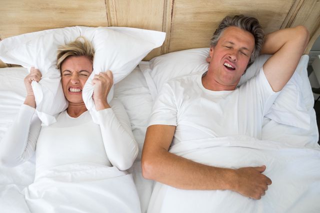 Annoyed woman covering ears with pillow while man snoring on bed - Download Free Stock Photos Pikwizard.com
