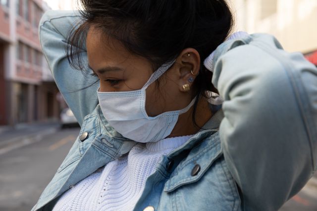 Close up of mixed race woman out and about in the city during the day, standing in the street and putting on a face mask for protection against coronavirus covid 19