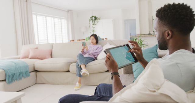 Diverse couple sitting on couch and using tablet in living room. Spending quality time at home concept.