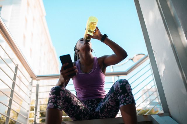 Fit african american woman resting on stairs, drinking water and using smartphone - Download Free Stock Photos Pikwizard.com