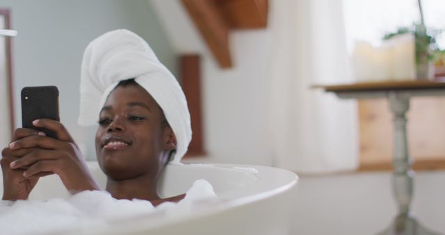 Smiling african american attractive woman relaxing in bath and using smartphone in bathroom. beauty, pampering, home spa and wellbeing concept.