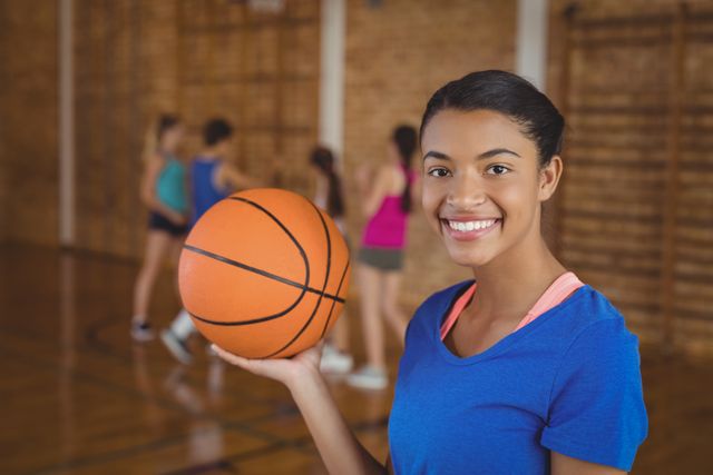 Portrait of smiling high school girl holding a basketball while team playing in background