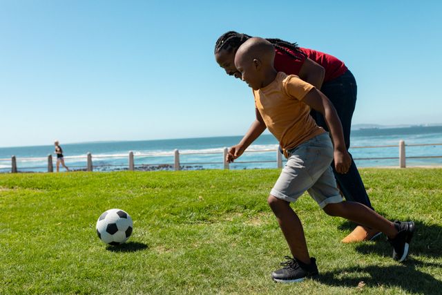 Full length of african american son playing soccer with father at grass promenade at beach. unaltered, enjoyment, family, lifestyle and togetherness concept.