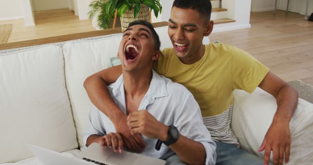 Smiling mixed race gay male couple sitting on sofa using laptop and laughing - Download Free Stock Photos Pikwizard.com