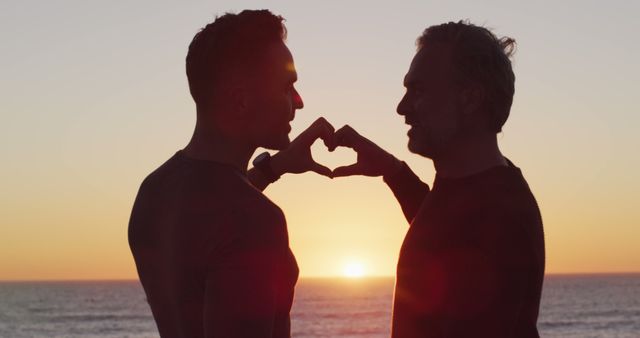 Happy caucasian gay male couple holding hands and making heart shape at sunset on the beach - Download Free Stock Photos Pikwizard.com