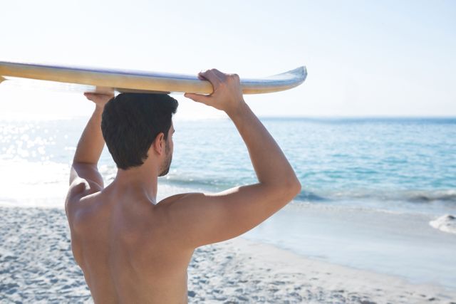 Rear view of shirtless man carrying surfboard at beach - Download Free Stock Photos Pikwizard.com